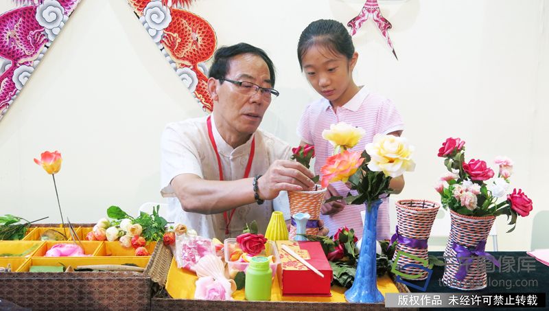 01 7月27日，第六届北京文学艺术品展示会在北京展览馆开幕。北京绢花代表性传承人金铁铃现场带领小朋友制作北京绢花、插花。千龙网记者 张嘉玉摄